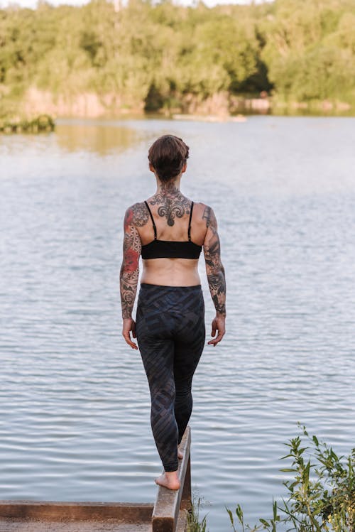 Back view of anonymous barefoot female in sportswear standing on wooden dock against lake and trees
