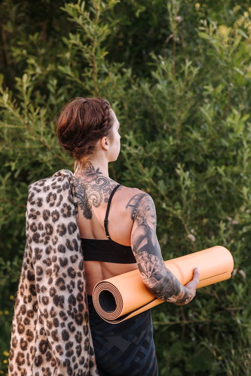 Anonymous tattooed woman with yoga mat and plaid near tree