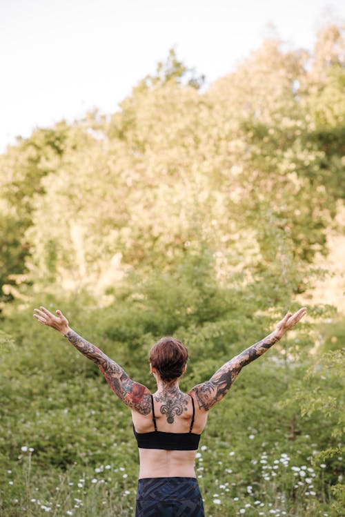 Unrecognizable tattooed woman practicing yoga against green trees