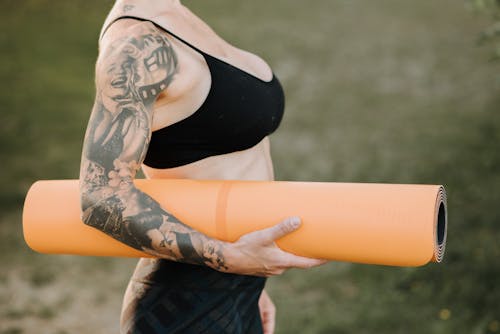 Free Side view of crop anonymous tattooed female in sports clothes with rolled yoga mat standing on lawn Stock Photo