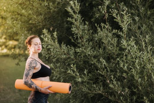 Side view of young slim contemplative female in sportswear with rolled yoga mat looking down in summer