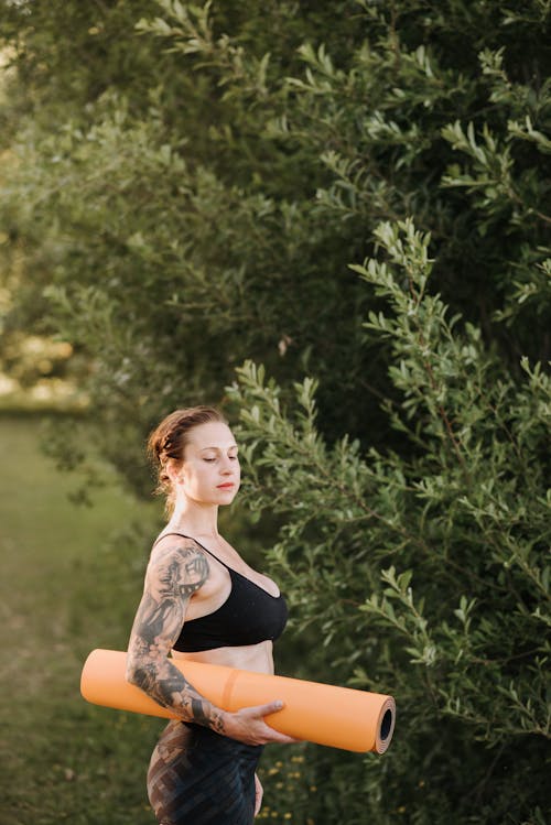 在绿色的树旁思考瑜伽垫的纹身的女人