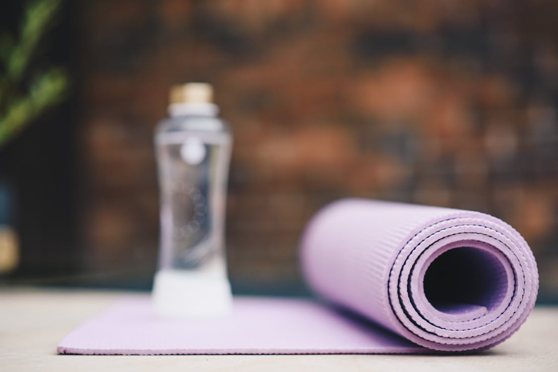 Roll of violet yoga mat with transparent plastic bottle of water on blurred background