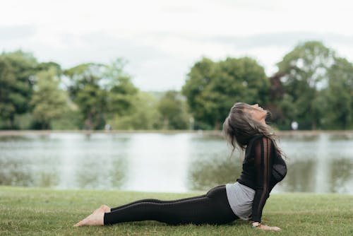 Side view of anonymous barefoot female in sportswear demonstrating High Cobra pose on grass coast against river