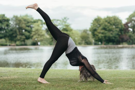 Side view of anonymous flexible female in sportswear standing in Eka Pada Adho Mukha Svanasana pose while practicing yoga on river coast