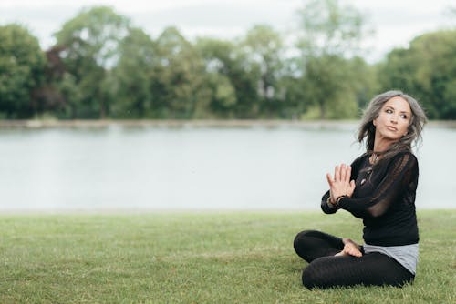 Side view of young flexible female in sportswear practicing yoga while sitting with crossed legs on river shore and looking away