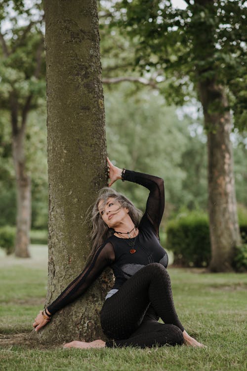 Young contemplative barefoot female in sports clothes sitting with crossed legs and raised arm on lawn near tree while practicing yoga and looking away