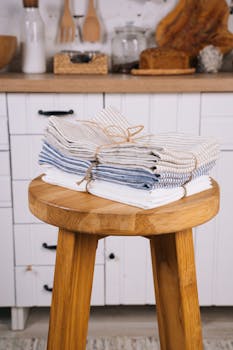A stack of linen kitchen towels neatly folded on a rustic wooden stool in a cozy kitchen setting.
