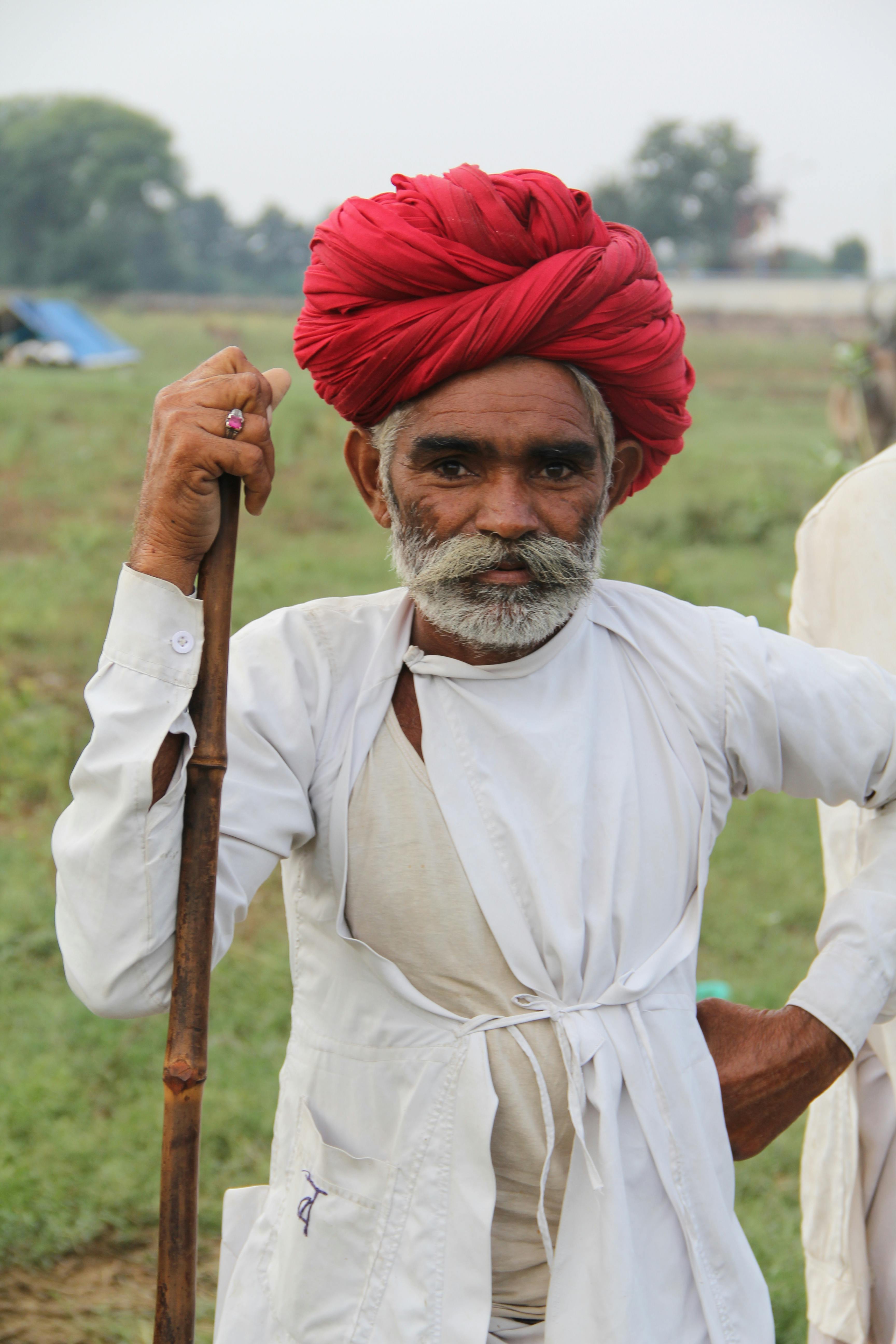 Elderly man with beard and turban poses outdoors in traditional attire holding a wooden staff.