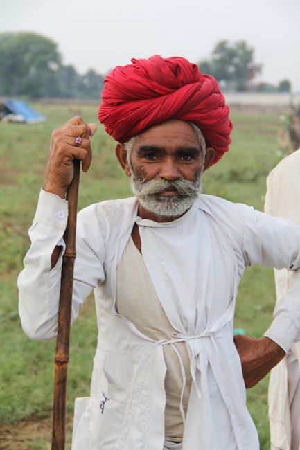 Elderly man with beard and turban poses outdoors in traditional attire holding a wooden staff.