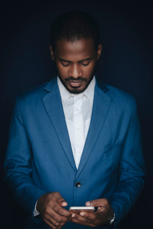 Free African American man in a blue suit focused on his smartphone, exuding professionalism. Stock Photo