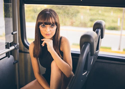 Young female in trendy glasses touching chin and looking at camera while sitting near window in modern bus