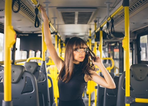 Young woman in trendy glasses touching hair and looking away while riding modern bus