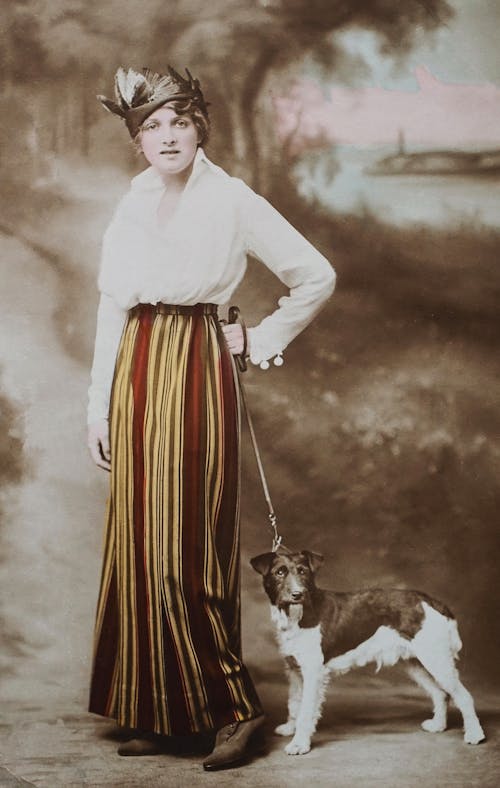 Woman in White Long Sleeve Shirt and Stripes Skirt Holding A Dog On Leash