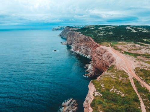Drone view of light blue cloudy sky above rocky formation covered with woodland and sandy roads next to cliffs over vibrant dark blue bay