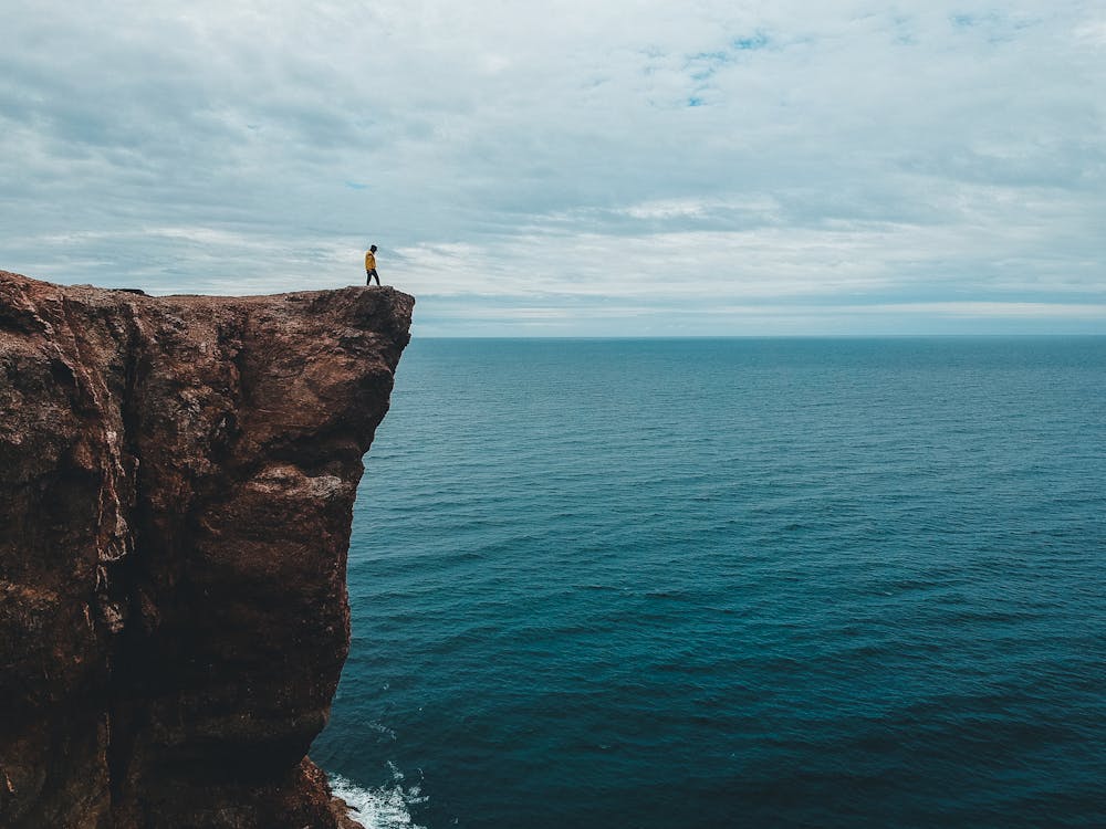 Grey overcast sky covered with clouds above tourist standing on edge of tall majestic dark brown cliff over wavy blue endless ocean