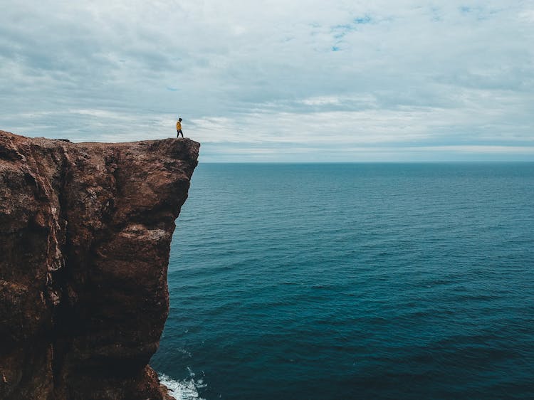 Faceless Hiker On Steep Rock Over Bay Under Overcast Sky