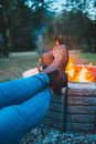 Unrecognizable tourist in jeans and boots chilling with legs crossed by bright bonfire in big fire bowl in forest on blurred background