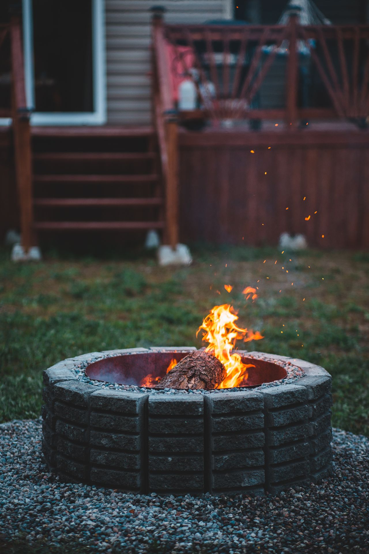 Fire pit with flames on a gravel patio area beside a deck
