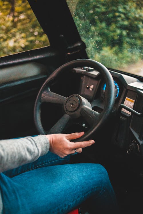Crop driver in casual clothing holding black round steering wheel while driving tractor on road surrounded by trees in daylight