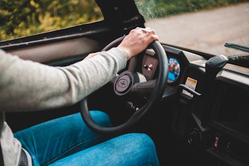 Faceless driver in grey sweater and jeans using big black round spinning wheel while driving automobile in countryside