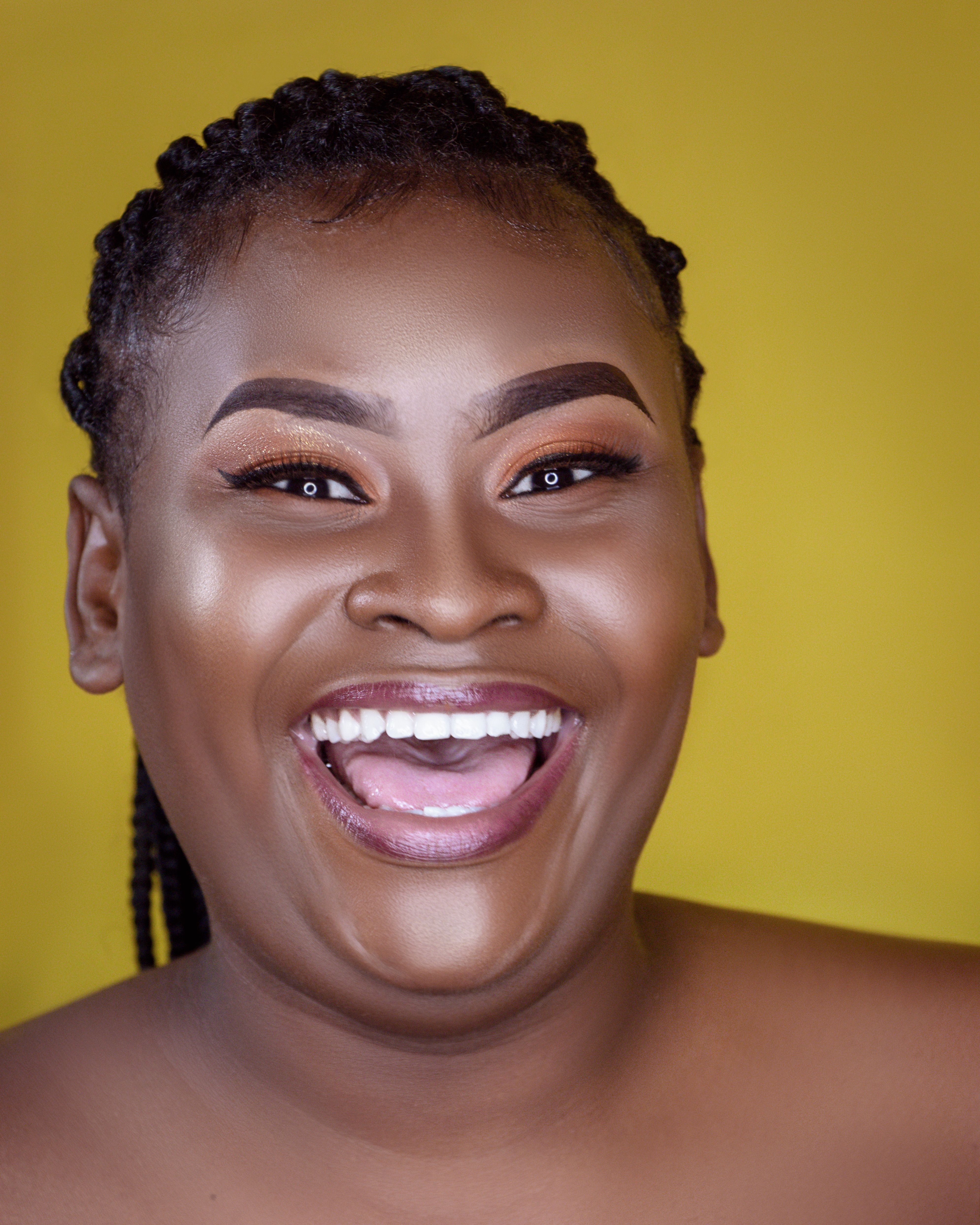 Close-up of a happy black woman with natural makeup and braided hair against a yellow background.