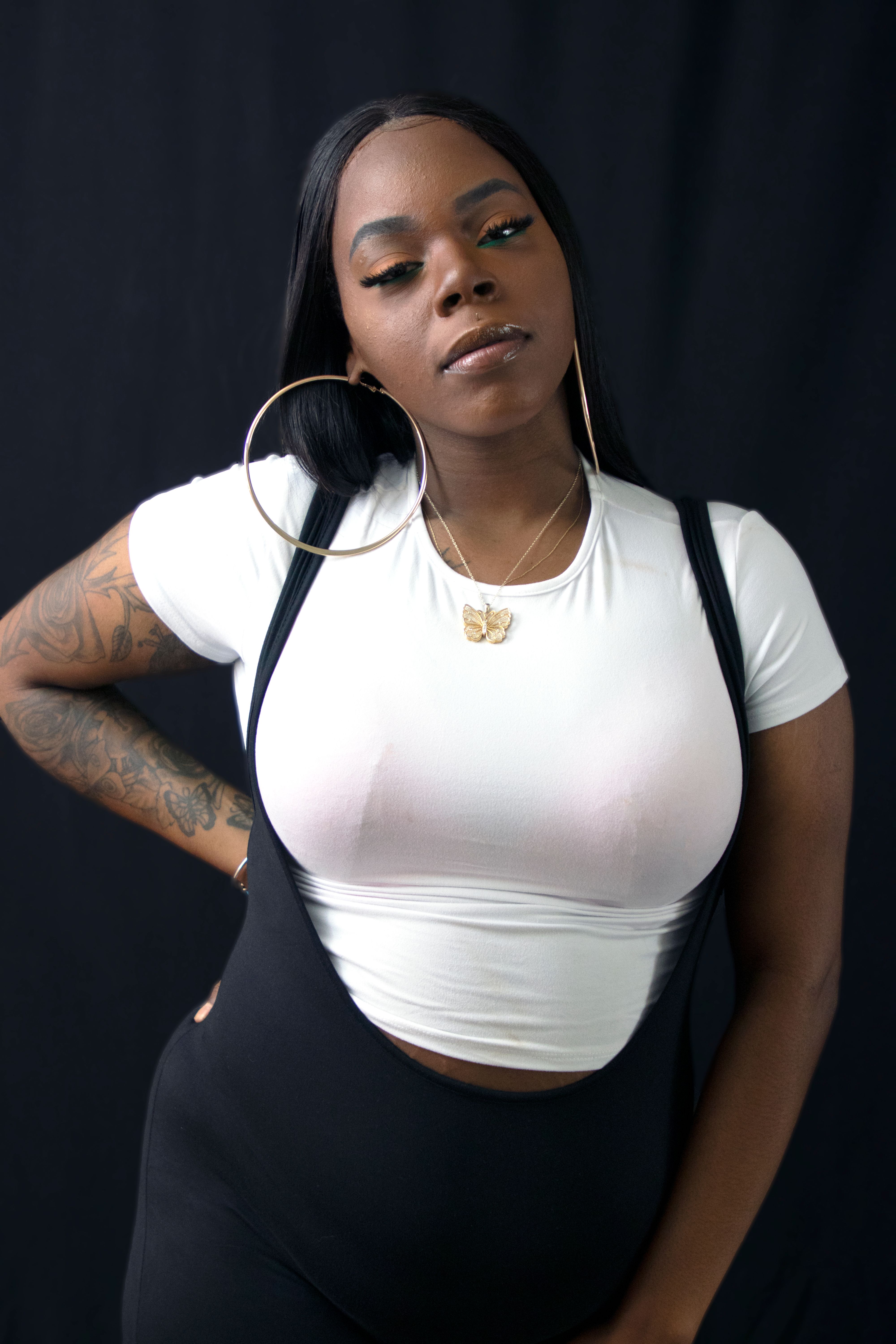 A confident plus-size woman posing stylishly with hoop earrings and a white shirt, showcasing her arm tattoo.