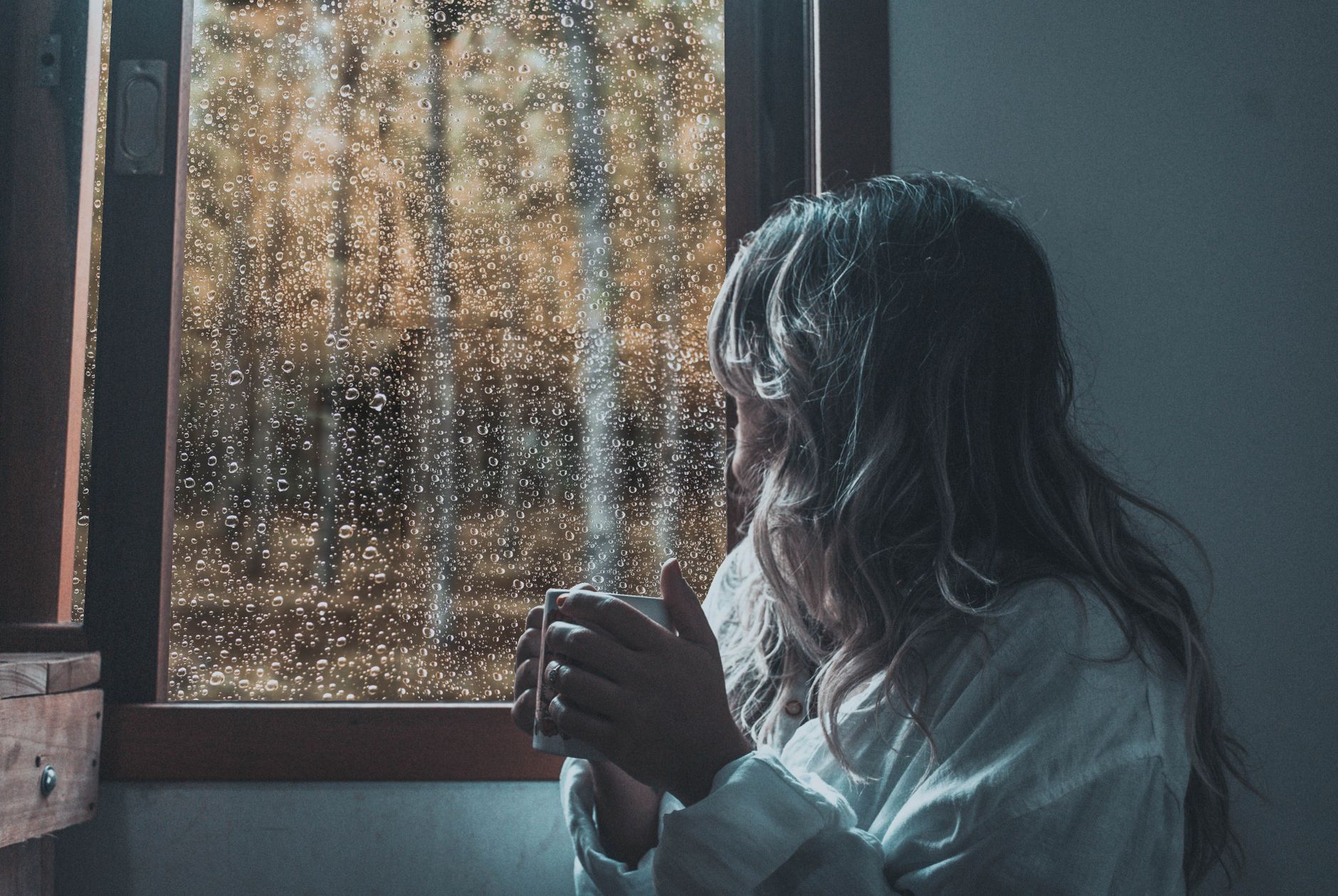 A woman in white sleeves holds a cup, gazing out a rain-soaked window.