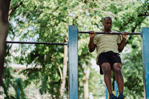An athletic adult man doing a pull-up on an outdoor metal bar surrounded by green trees.