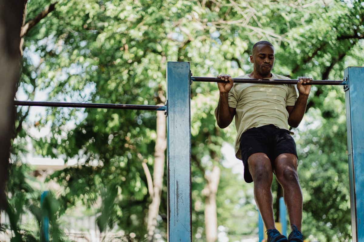A man doing a pull-up on an outdoor metal bar in a park surrounded by trees.