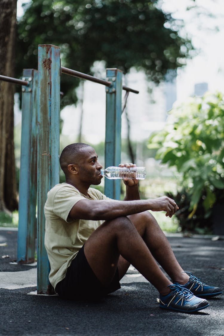 A Tired Man Sitting On The Ground While Drinking Water On A Plastic Bottle