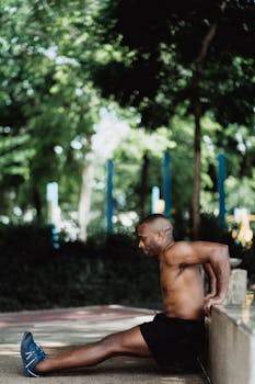 Topless man exercising outdoors, leaning against a wall in a park setting for fitness routine.
