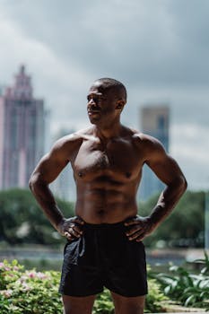 Shirtless muscular man standing confidently outdoors, showcasing fitness and strength with urban backdrop.