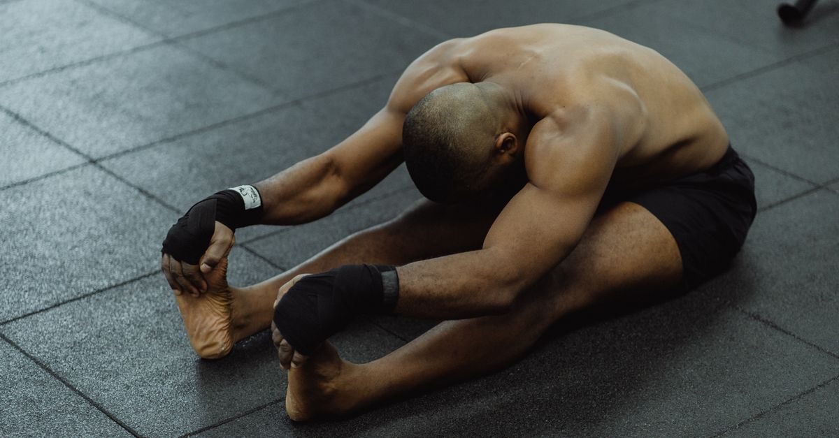 A shirtless man stretches on a gym floor, barefoot and wearing black shorts, focused on flexibility.