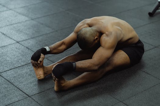 A shirtless man stretches on a gym floor, barefoot and wearing black shorts, focused on flexibility.