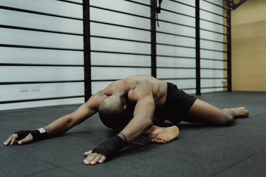 A shirtless man stretches on a black mat in an indoor gym with metal bars.