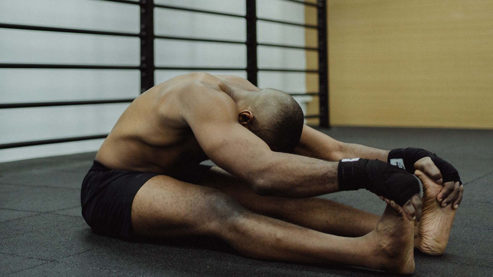 Topless man sitting on gym floor, stretching, showcasing fitness and flexibility.