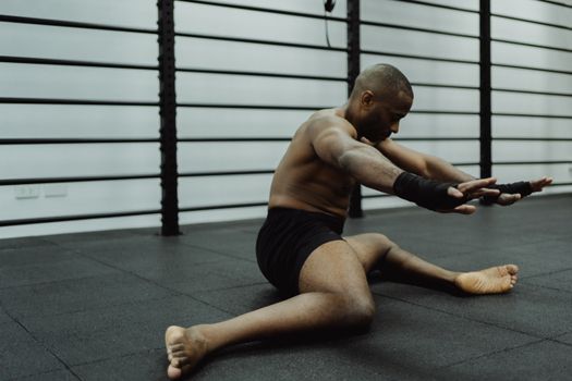 Topless man with hand wraps stretching on gym mat, preparing for workout.