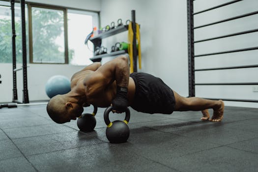 A shirtless man with a muscular build exercises in a gym, performing push-ups using kettlebells.