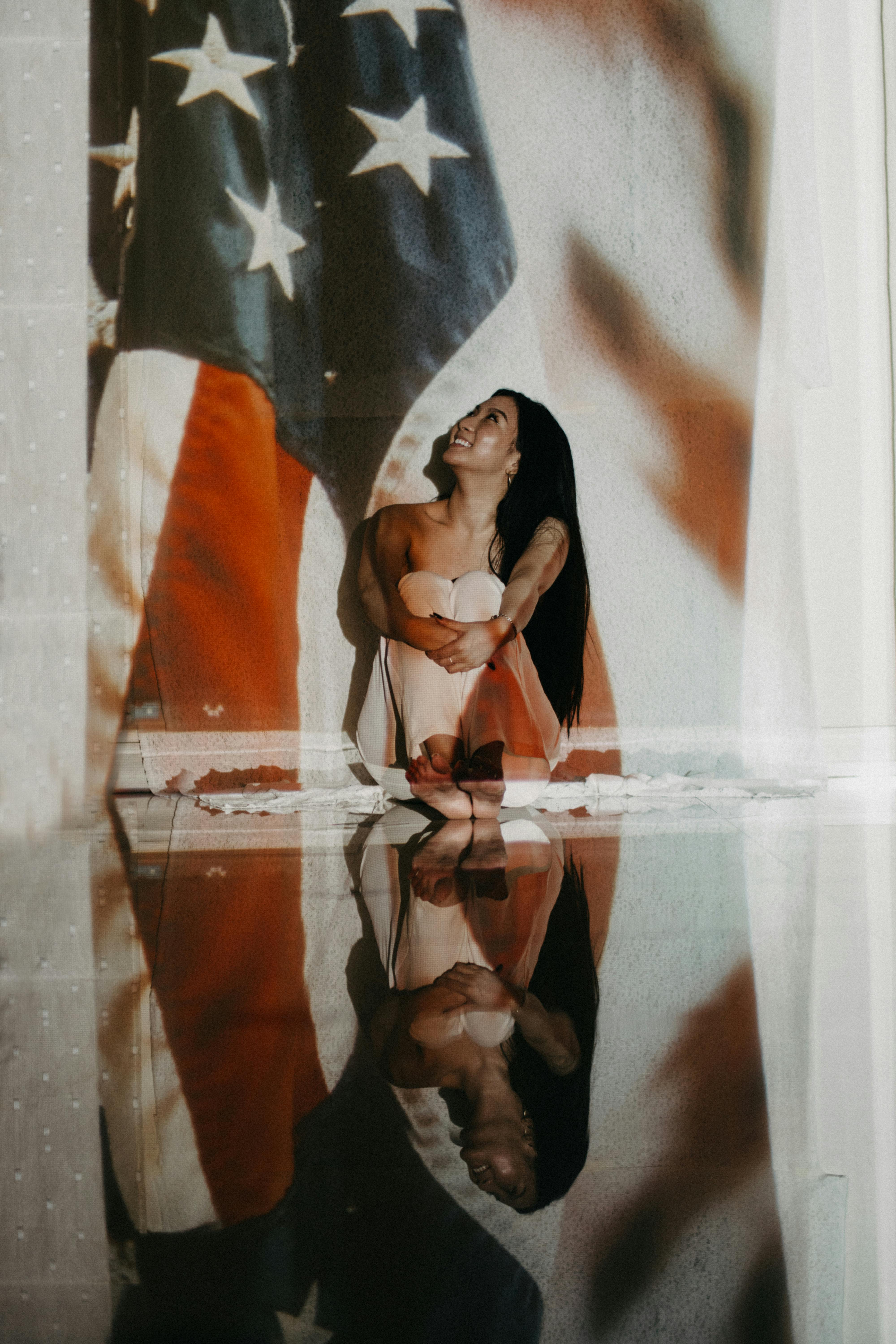 A woman sitting in front of a large American flag with her reflection visible below.