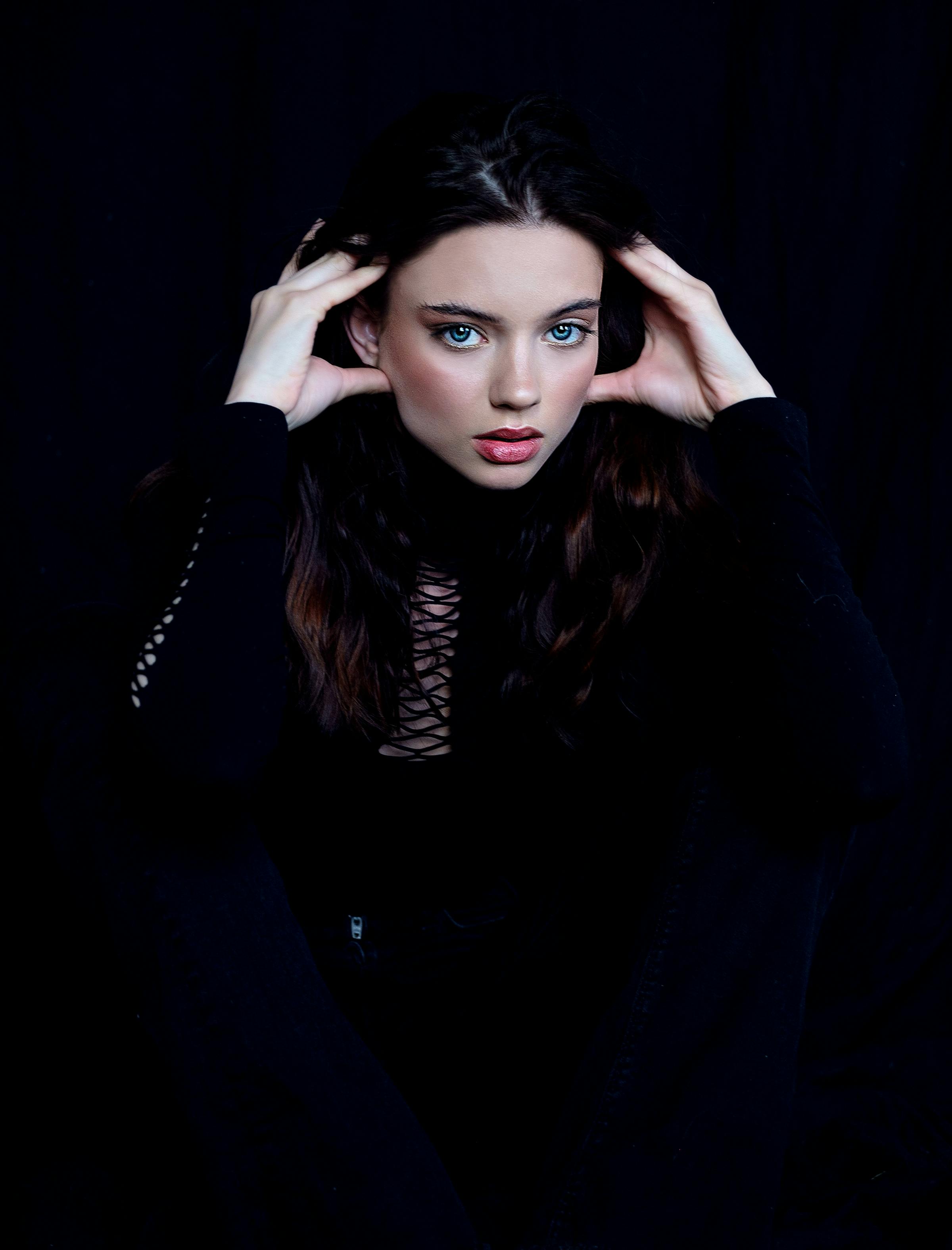Beautiful young female in black clothes touching head tenderly and looking at camera during photo session in dark studio