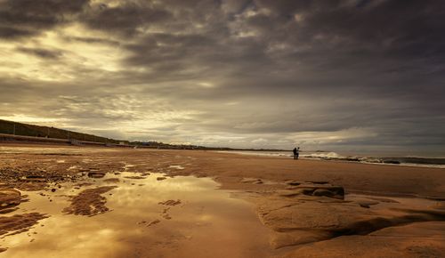 Free A dramatic sunset over a tranquil beach with reflections and a lone figure in the distance. Stock Photo
