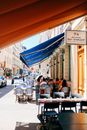 People sitting on chairs at tables on terrace of cafeteria with marquise on sunny sunny street with cars in city