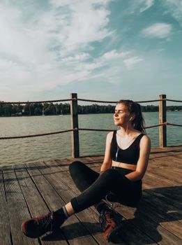Relaxing on a lakeside pier, a woman in sportswear enjoys the sunlight with earphones.