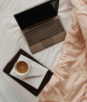 Overhead view of netbook near diary with paper and mug of hot drink on crumpled bed sheet