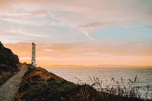 Beacon on mount with narrow path near rippled sea under bright cloudy sky at sunset