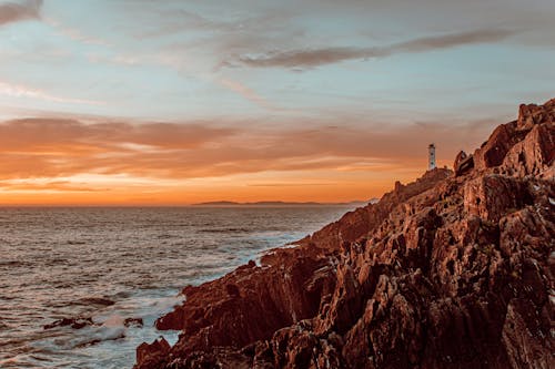 Amazing scenery of lighthouse in distance located near rocky coast with waving ocean with sun setting on horizon in summer evening