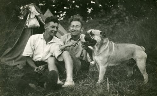 Grayscale Photo of Young Men Sitting Beside a Bulldog
