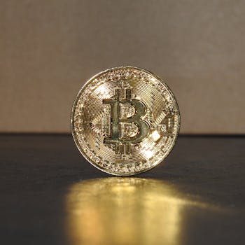 A shimmering golden Bitcoin coin standing upright on a reflective surface, illuminated by warm light.