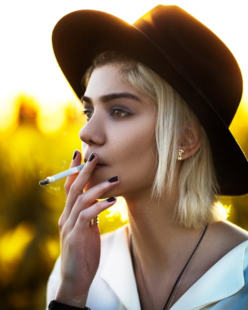 Young dreamy female in trendy apparel and felt hat smoking cigarette while looking away